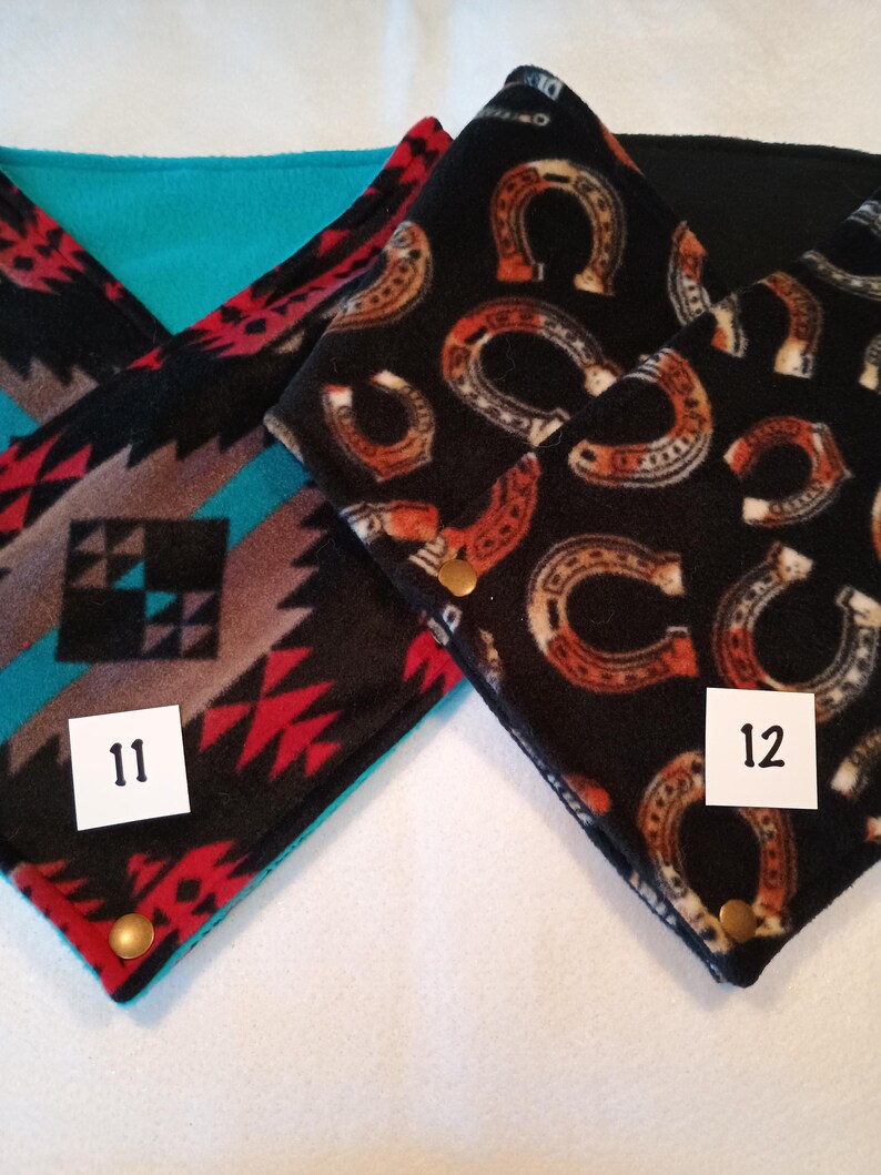 K&ouml;nnte beinhalten: Zwei Fleece-Halsw&auml;rmer mit unterschiedlichen Mustern. Einer ist schwarz mit einem roten, wei&szlig;en und braunen geometrischen Muster und der andere ist schwarz mit einem braunen Hufeisenmuster. Beide haben ein t&uuml;rkisfarbenes Fleecefutter und einen goldenen Druckknopf. Die Halsw&auml;rmer sind mit den Nummern 11 und 12 gekennzeichnet.