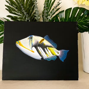 May include: A vibrant fish painting on a black canvas. The fish has a white body with yellow, blue, and black markings. The artwork is set against a backdrop of green foliage and a white vase.