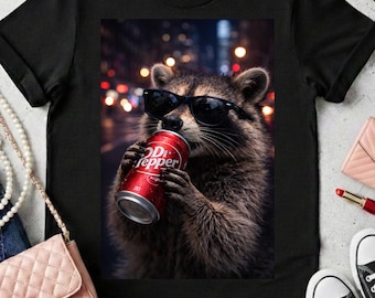 May include: Black t-shirt featuring a raccoon wearing sunglasses and drinking from a Dr. Pepper can. The raccoon is set against a blurred city lights background. Accessories include sunglasses, jewelry, and a purse.