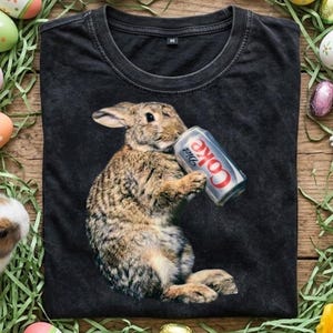 Può includere: T-shirt nero con un coniglio che beve da una lattina di Diet Coke. Il coniglio è marrone e bianco, con la lattina di Diet Coke in argento, rosso e bianco. La maglietta ha un girocollo e maniche corte. Uova di Pasqua ed erba sullo sfondo.