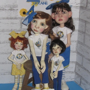 May include: Three dolls wearing colorful outfits and holding peace signs. The dolls are standing in front of a white brick wall with a sign that says "Pray for Ukraine".