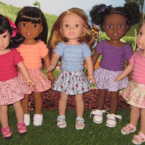 May include: Five dolls wearing different colored knitted tops and patterned skirts. The dolls are standing on a green grassy surface.