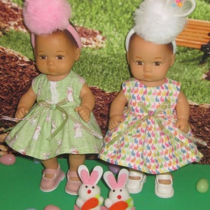 May include: Two dolls wearing Easter bunny headbands and dresses with bunny patterns. The dolls are holding bunny-shaped plush toys with carrots in their mouths.
