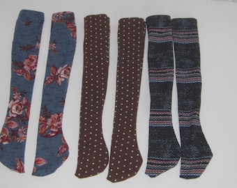 May include: Six pairs of fabric doll leggings in various patterns. Two pairs are blue with floral designs, two pairs are brown with white polka dots, and two pairs are blue with a striped pattern.