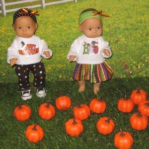 May include: Two baby dolls dressed in Halloween costumes, one wearing a white shirt with "Happy Halloween" and black and white polka dot pants, the other wearing a white shirt with "Boo" and a colorful striped skirt. The dolls are surrounded by 12 small orange pumpkins.