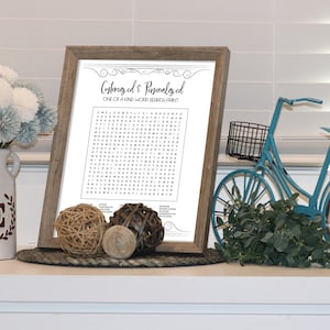 May include: A framed word search puzzle with the title "Customized & Personalized One of a Kind Word Search Paint". The puzzle is printed on white paper with black text. The frame is brown wood with a white mat. The puzzle is sitting on a white shelf with a blue bicycle, a clock, and a vase of flowers.