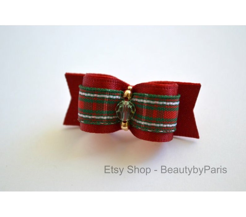 May include: A red, green, and white plaid ribbon bow with a clear crystal accent. The bow is attached to a red felt backing.