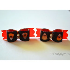 May include: Two black and orange Halloween-themed hair bows with silver beads. Each bow features a different design: a slice of pumpkin pie and a candy corn ghost.