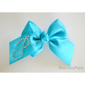 May include: A bright turquoise satin bow with a rhinestone-studded letter "B" on one tail. The bow is tied in a classic style, with a knot in the center and symmetrical loops. The bow is a hair accessory.