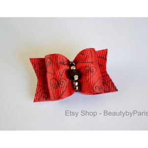 May include: A red bow with a black and silver bead in the center. The bow has a black swirl pattern on it. Etsy Shop - BeautybyParis.