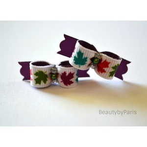 May include: Two white hair bows with a fall leaf pattern in red, green, and orange. The bows are decorated with small, iridescent beads.