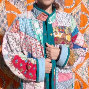 May include: A colourful patchwork jacket featuring floral and patterned fabrics in red, blue, green, and yellow. The jacket has teal trim, light blue cuffs, and gold buttons. It is a quilted jacket.