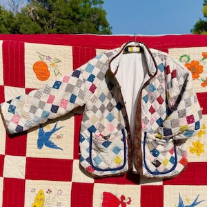 May include: A quilted jacket with a patchwork design in various colors, including blue, pink, and white. The jacket has two pockets and a brown trim. It is hanging on a hanger against a red and white quilt background.