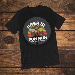 Puede incluir: Una camiseta negra de manga corta con un gráfico circular sobre un fondo de atardecer retro. El diseño incluye siluetas de personas corriendo, un platillo volante y el texto AREA 51 FUN RUN SEPTEMBER 20 2019 en letras de color blanco.