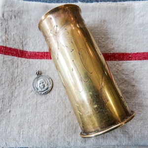 May include: A cylindrical brass shell casing featuring etched floral designs and a small circular silver-toned metal pendant depicting a religious figure in profile. The brass object has a smooth, metallic finish with visible engravings.