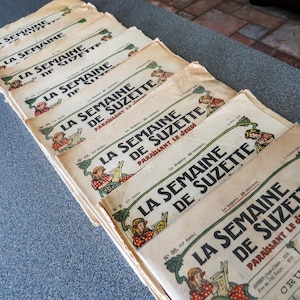 Może przedstawiać: Stos starych francuskich magazynów zatytułowanych "La Semaine de Suzette" jest wystawiony. Okładki zawierają ilustracje dziewczynki czytającej i dekoracyjne zielone winorośle. Magazyny mają postarzaną, pożółkłą papierową fakturę.