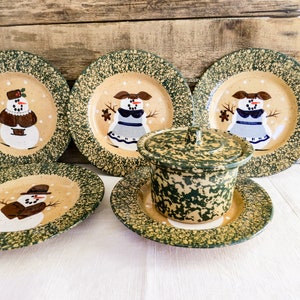Puede incluir: Conjunto de cinco platos de cerámica moteados en verde y beige con diseños de muñeco de nieve. Los platos tienen un borde verde y un centro blanco con un muñeco de nieve pintado en marrón, negro, blanco y rojo. El conjunto también incluye un plato con tapa de cerámica moteado en verde y beige a juego.