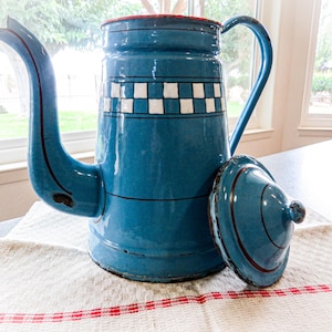 Puede incluir: Una cafetera alta de esmalte azul con un pico largo y curvado y una tapa a juego colocada al lado. La cafetera presenta una banda horizontal de cuadrados a cuadros blancos y azules cerca de la parte superior, acentuada por finas líneas negras.