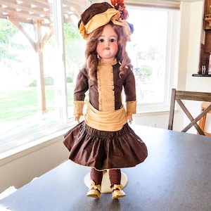 May include: A vintage doll with brown hair and a painted face, wearing a brown dress with a tan bodice and a matching hat. The doll has gold shoes and is standing on a white stand.