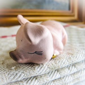 May include: A small pink ceramic pig figurine with black painted eyes and eyelashes, lying on its side with its head resting on a folded linen fabric.