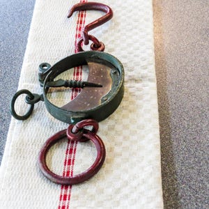 Könnte beinhalten: Eine antike Waage mit einem kreisförmigen Metallrahmen, einer gebogenen Klinge und einem roten Haken und Ring. Die Waage liegt auf einem weiß-rot gestreiften Tuch.