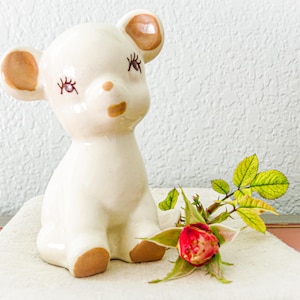 May include: A white ceramic figurine of a fawn with brown ears and eyes. The fawn is sitting on a white surface with a single red rose and green leaves.