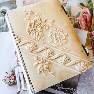 Puede incluir: Álbum de fotos antiguo de color crema con detalles florales y monogramas en relieve. El álbum presenta un borde decorativo y está rodeado de tarjetas religiosas vintage y un textil a rayas.