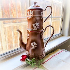May include: A brown enamel coffee pot with white floral designs. The pot has two sections, a top section for brewing and a bottom section for holding the coffee. The pot is sitting on a white surface with a red rose.