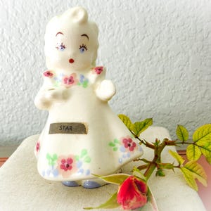Puede incluir: Una figurita de porcelana blanca de una niña con un vestido floral. La figurita tiene ojos azules y boca rosa. El vestido tiene flores rosas, azules y verdes. La figurita está etiquetada con "STAR".