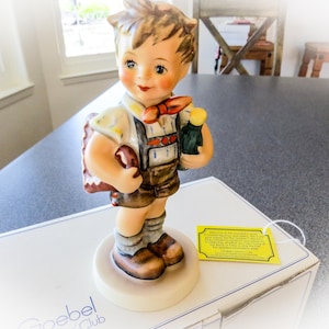 May include: A porcelain figurine of a young boy in traditional German attire, holding a satchel and a small bottle. The figurine has a detailed painted design with a bow tie and is standing on a white base. The figurine is on top of a white box.