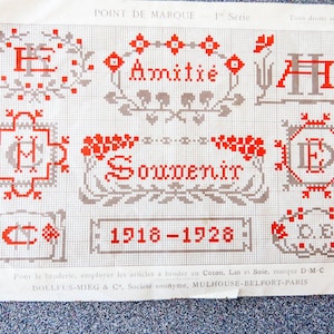 Könnte beinhalten: Ein Vintage-Kreuzstichmusterbogen mit roten und grauen Designs auf einem Rasterhintergrund. Das Blatt enthält die Wörter "Amitié" und "Souvenir" sowie die Jahre "1918-1928". Die Designs umfassen florale und geometrische Motive.
