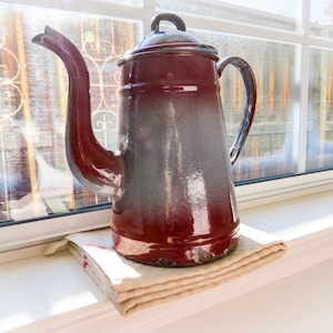 May include: A red and gray enamel teapot with a handle and spout. The teapot is sitting on a white linen napkin on a white windowsill.