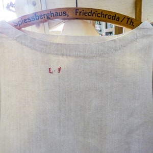 May include: A light-colored linen shirt with a simple round neckline. The shirt has short sleeves and a small embroidered detail in red thread near the neckline. The shirt is hanging on a wooden hanger.