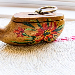 May include: A wooden trinket box shaped like a clog with a hinged lid and a metal ring handle. The box is painted with red, yellow, and green flowers.