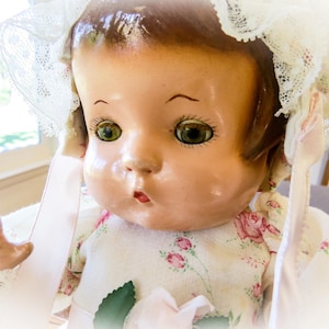 Op de afbeelding: Een vintage pop met een geschilderd gezicht, groene ogen en een kleine rode mond. De pop draagt een bloemenjurk en een kanten muts. De huid van de pop heeft een glanzende afwerking en de algehele esthetiek doet denken aan antiek speelgoed.