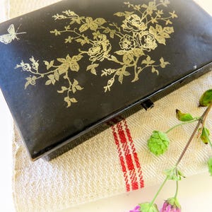 May include: A black rectangular box with a gold floral design and a butterfly. The box is resting on a beige and red striped cloth, with a sprig of greenery and purple flowers. The box has a small latch.