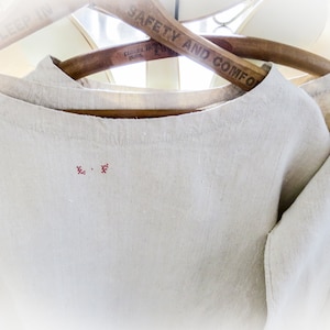 May include: Off-white linen top with a round neckline, hanging on a wooden hanger. The top features a small, embroidered red design near the neckline. The hanger reads "SAFETY AND COMFORT".