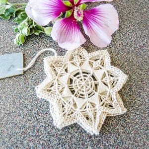 May include: A cream-colored, star-shaped doily with intricate crochet details. A small, light blue tag is attached with a string. A pink and purple flower with green leaves is in the background. The doily is on a speckled gray surface.