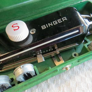 May include: A vintage Singer sewing machine in a green case. The black machine has a white knob with a red "S" and the Singer logo in white. The case contains silver metal bobbins and other parts.