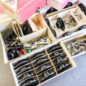 Puede incluir: Accesorios y piezas de máquinas de coser Singer vintage organizados en cajas de cartón abiertas. Las cajas están etiquetadas con el logotipo de Singer. El contenido incluye piezas metálicas negras, bobinas y varios accesorios de máquinas de coser.