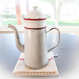 May include: A vintage speckled white coffee pot with a red trim on the lid and base, featuring a curved spout and handle. It rests on a folded white cloth with red stripes, suggesting a rustic kitchen theme.
