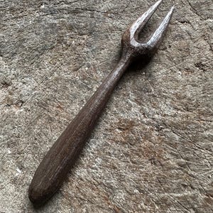 May include: A rustic, two-pronged fork with a dark brown, textured handle. The fork's tines are slightly curved and pointed. The utensil is made of wood and rests on a weathered, gray wooden surface. The overall aesthetic is reminiscent of handcrafted kitchenware.