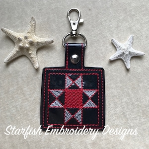 May include: A black, red, and silver embroidered quilt block keychain with a silver metal clasp. The keychain is attached to a black leather strap with a silver snap button. The text "Starfish Embroidery Designs" is visible at the bottom of the image.