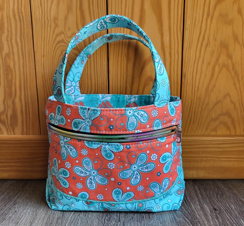 May include: A small turquoise and orange fabric tote bag with a zipper closure. The bag has a floral pattern and is perfect for carrying small items.