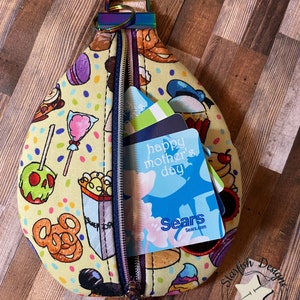 May include: A yellow raindrop-shaped key fob pouch with a zipper closure and a colourful print of various Disney-themed food items. The pouch is open and has a blue and white gift card with the text "happy mother's day" and a white gift card with the text "Sears" inside.