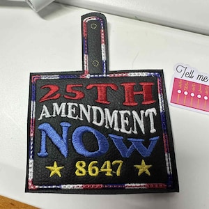 May include: Black leather patch with the text "25TH AMENDMENT NOW" in red and blue. The patch has a rectangular shape with a small handle at the top. The number "8647" and two yellow stars are also visible.