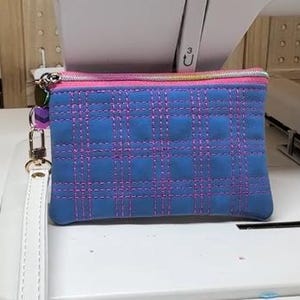 May include: A blue quilted pouch with a plaid pattern stitched in pink thread. The pouch has a pink zip and a white wrist strap with a gold-coloured clasp. The pouch is on a white surface.