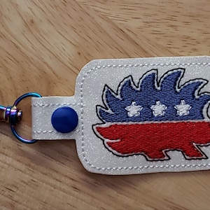May include: A white glitter keychain with a blue and red embroidered porcupine design. The porcupine has three white stars on its back.