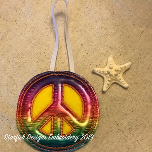 May include: A rainbow peace sign ornament with a yellow center and a white hanging loop. The ornament is embroidered with a peace sign and has a shiny, iridescent finish. The text "Starfish Designs Embroidery 2019" is visible at the bottom of the ornament.