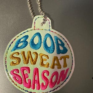 May include: A white, round ornament with a silver chain. The ornament is embroidered with the text "BOOB SWEAT SEASON" in colorful thread.